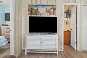 Ein Zimmer mit einem großen Flachbildfernseher, der an einem weißen Schrank befestigt ist, einem Strandbild darüber und einer Holztür, die in einen anderen Raum führt.