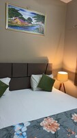 Una cama con ropa de cama blanca, almohadas verdes, colcha con estampado floral, un cuadro de una casa junto a un lago y una lámpara de pie.