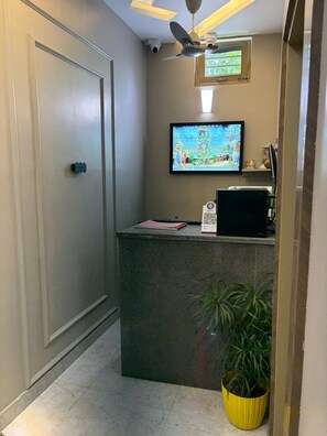 Un área de recepción con puerta cerrada, ventilador de techo, televisor montado, mostrador de recepción, planta en maceta y suelo de baldosas.