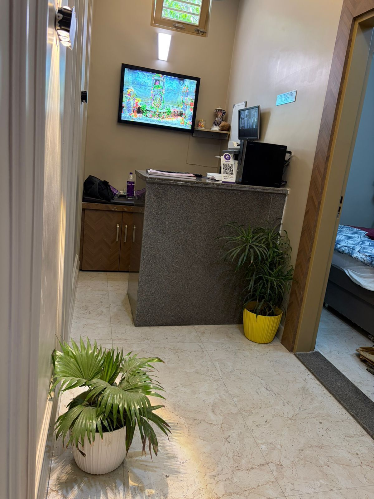 Ein Empfangsbereich mit einer Rezeption, einem an der Wand befestigten Fernseher, einer kleinen Vitrine und einer Topfpflanze.