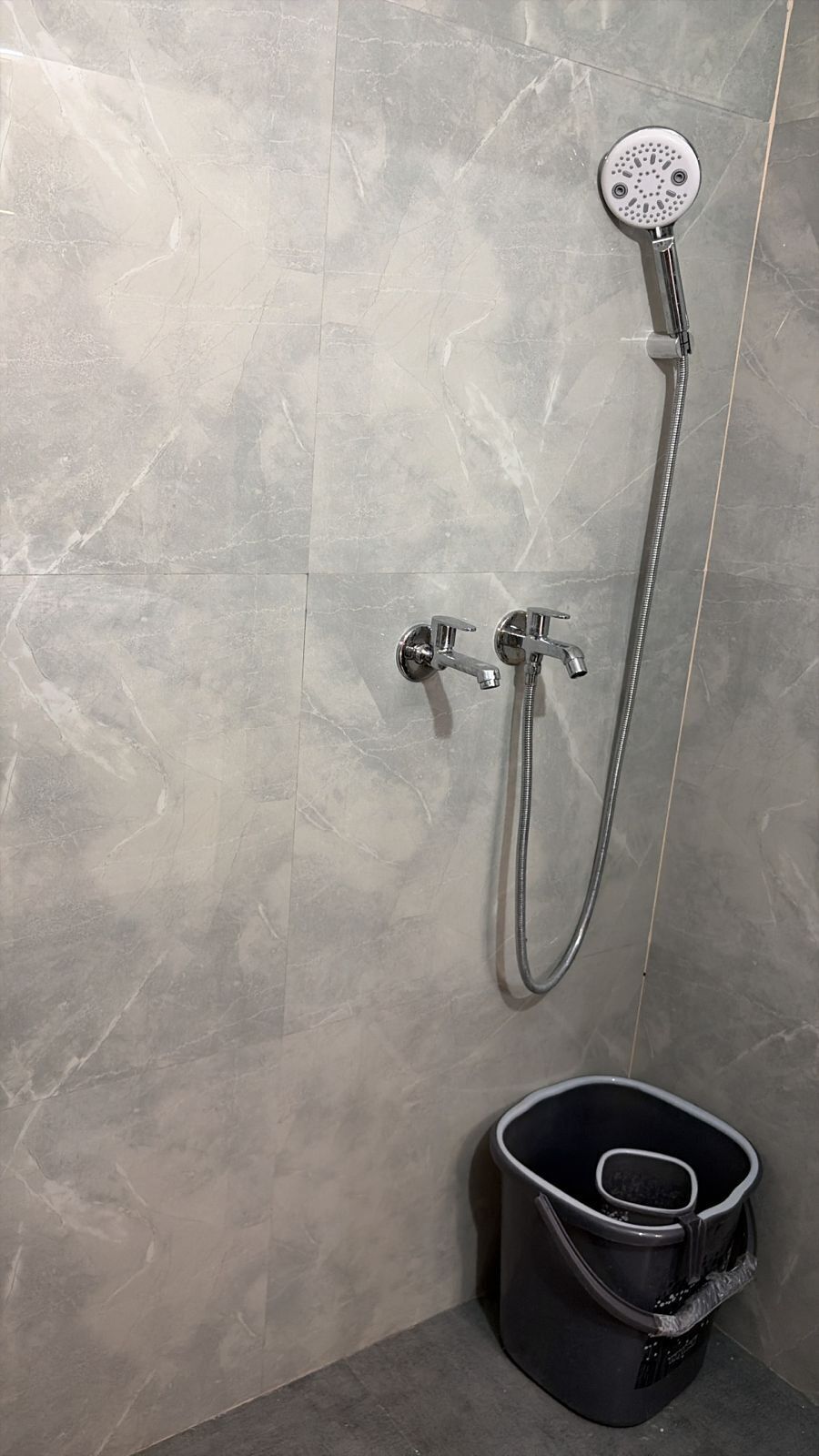 A shower with a handheld showerhead and a wall-mounted faucet, a black bucket, and a tiled wall.