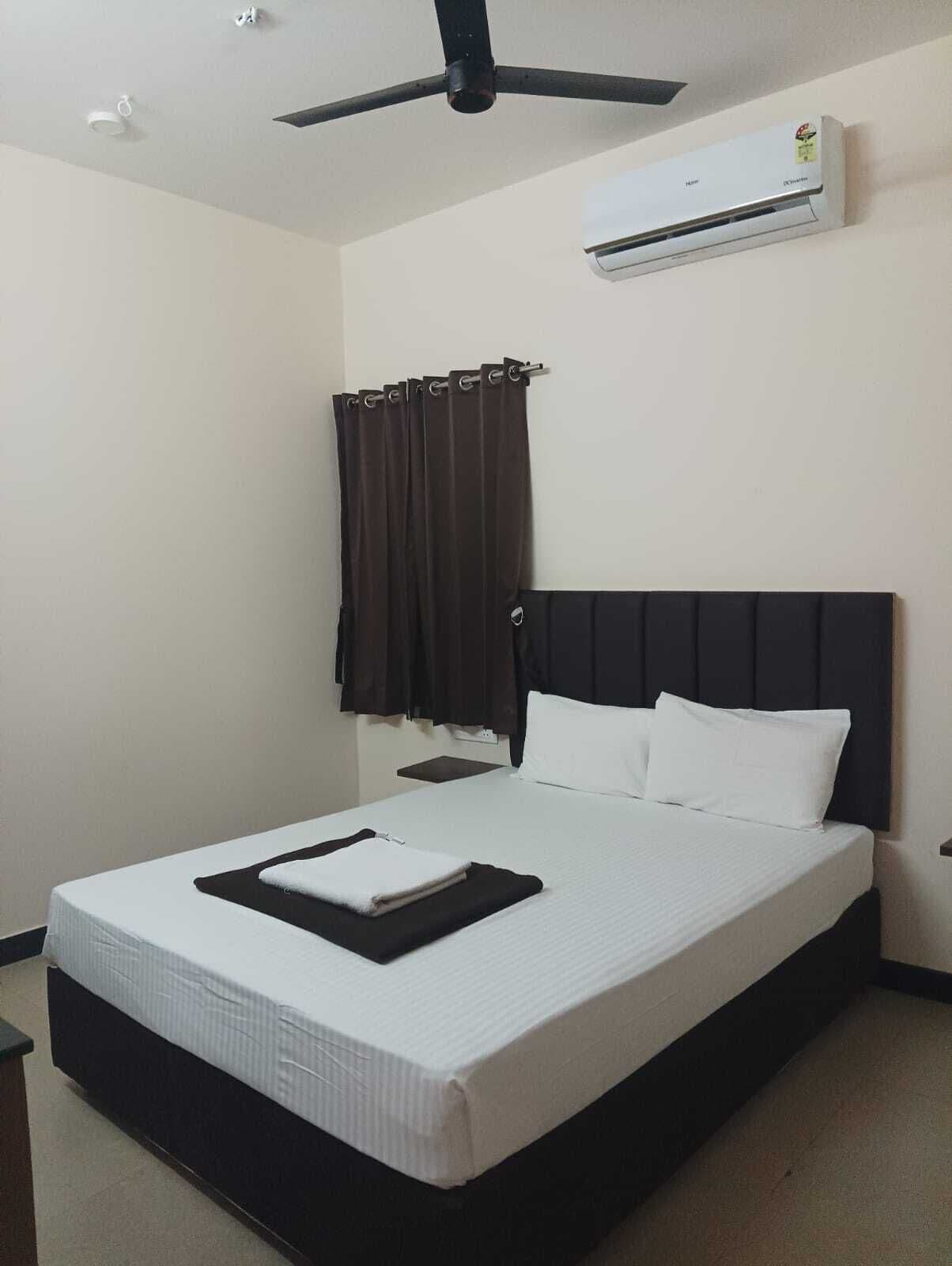 A hotel room with a bed, a ceiling fan, an air conditioning unit, and a window with curtains.
