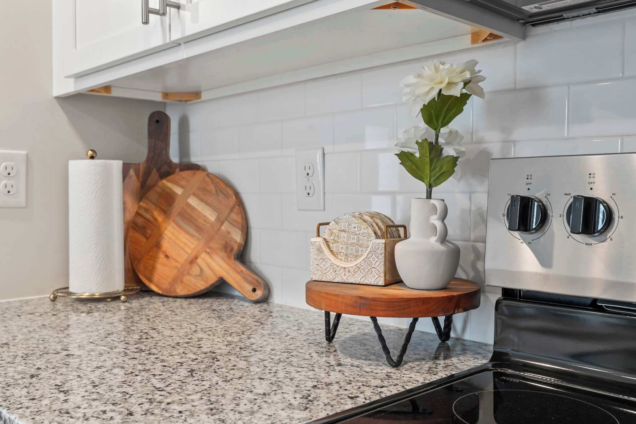 A kitchen with a granite countertop, a wooden cutting board, a white vase with flowers, a white kettle, and a stainless steel oven.