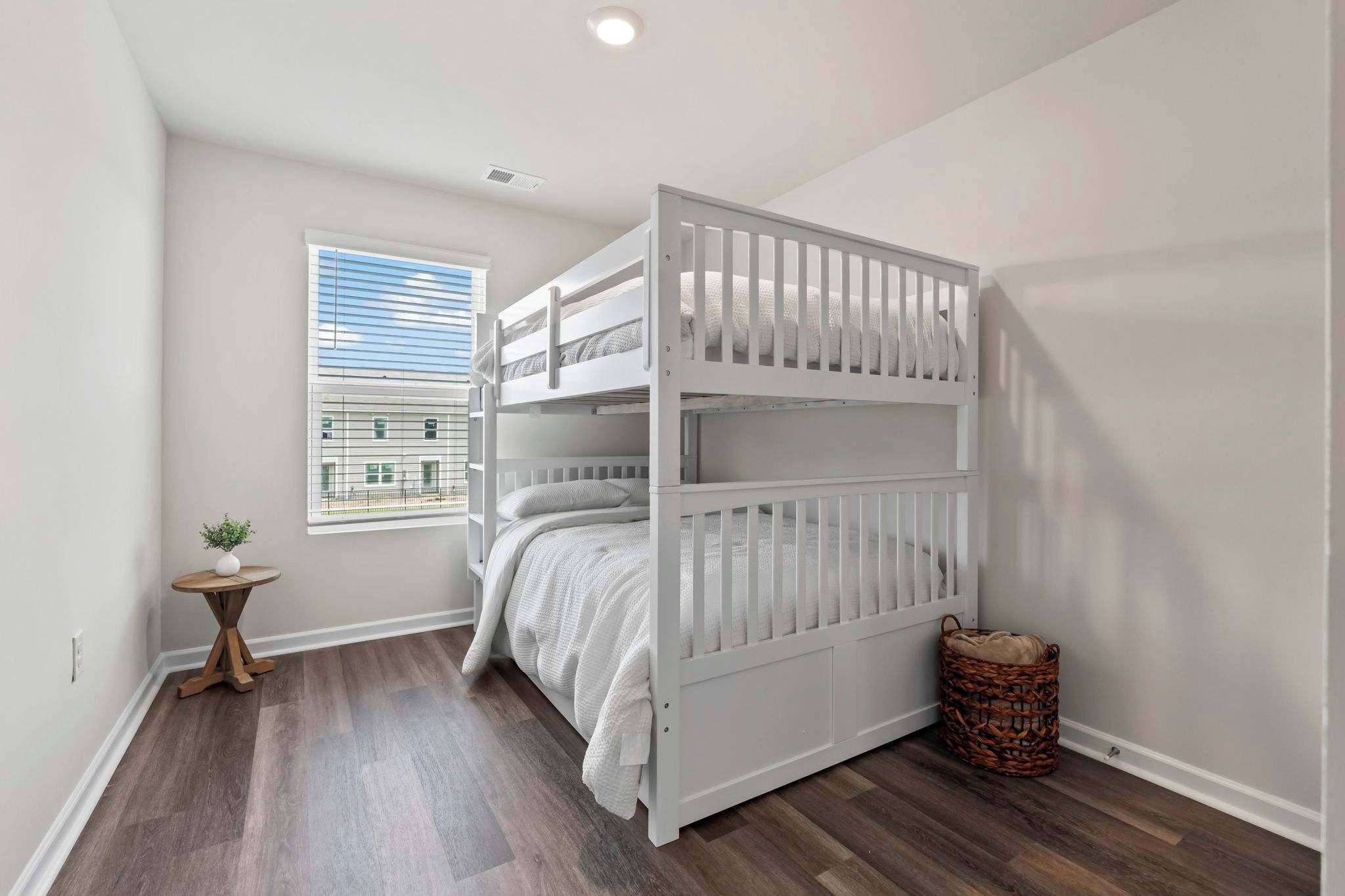 A bunk bed in a room with wooden flooring, a small table with a plant, a window with blinds, and a basket.