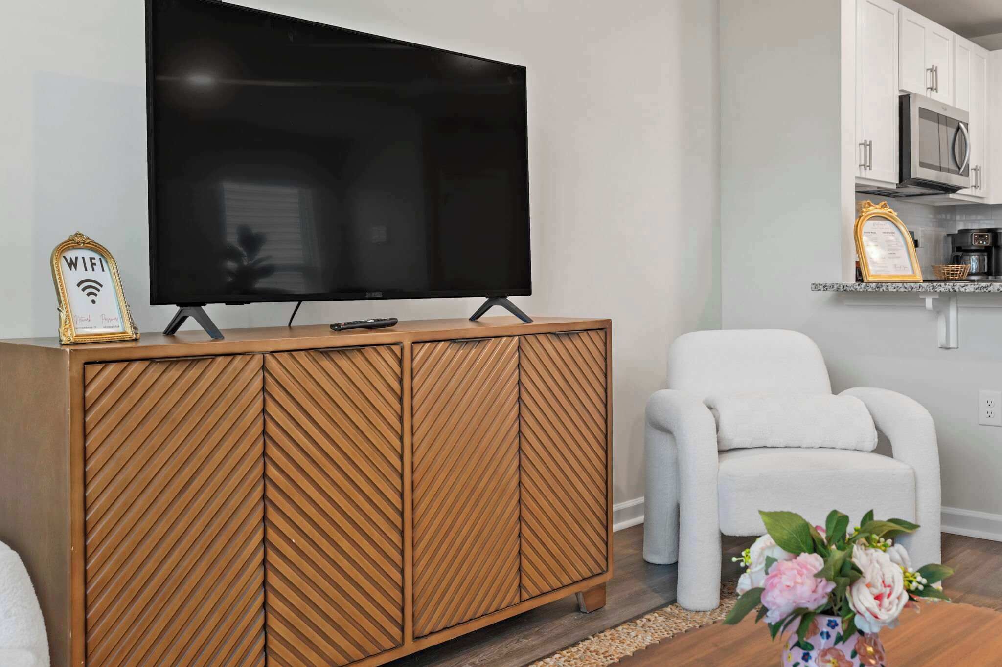 A modern living room with a large flat-screen TV, a wooden cabinet, a white armchair, and a small table with a vase of flowers.