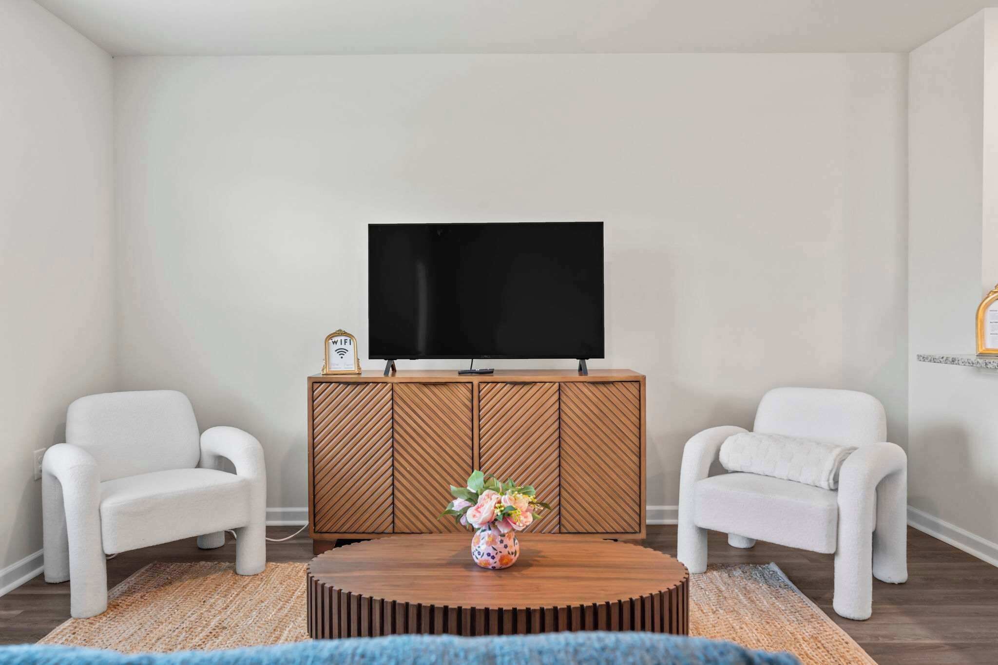 A living room with a TV on a wooden cabinet, two white armchairs, and a coffee table with a vase.
