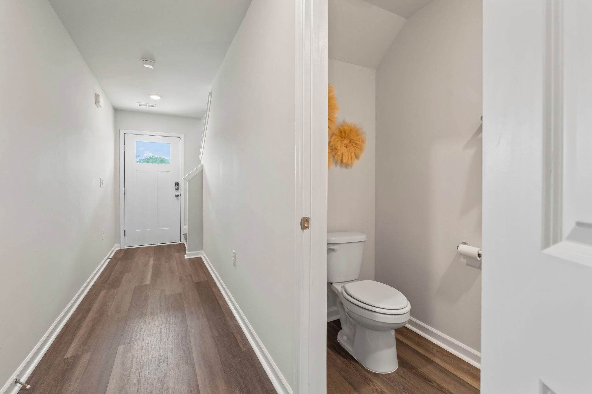 Ein Flur mit Holzboden, einer weißen Tür am Ende und einem weißen WC mit einem flauschigen Accessoire im Badezimmer.