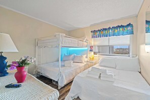 Ein Hotelzimmer mit Etagenbett, einem Einzelbett, einem Fenster mit Blick, einer Lampe, einer Vase mit Blumen und einem Tisch mit Telefon.