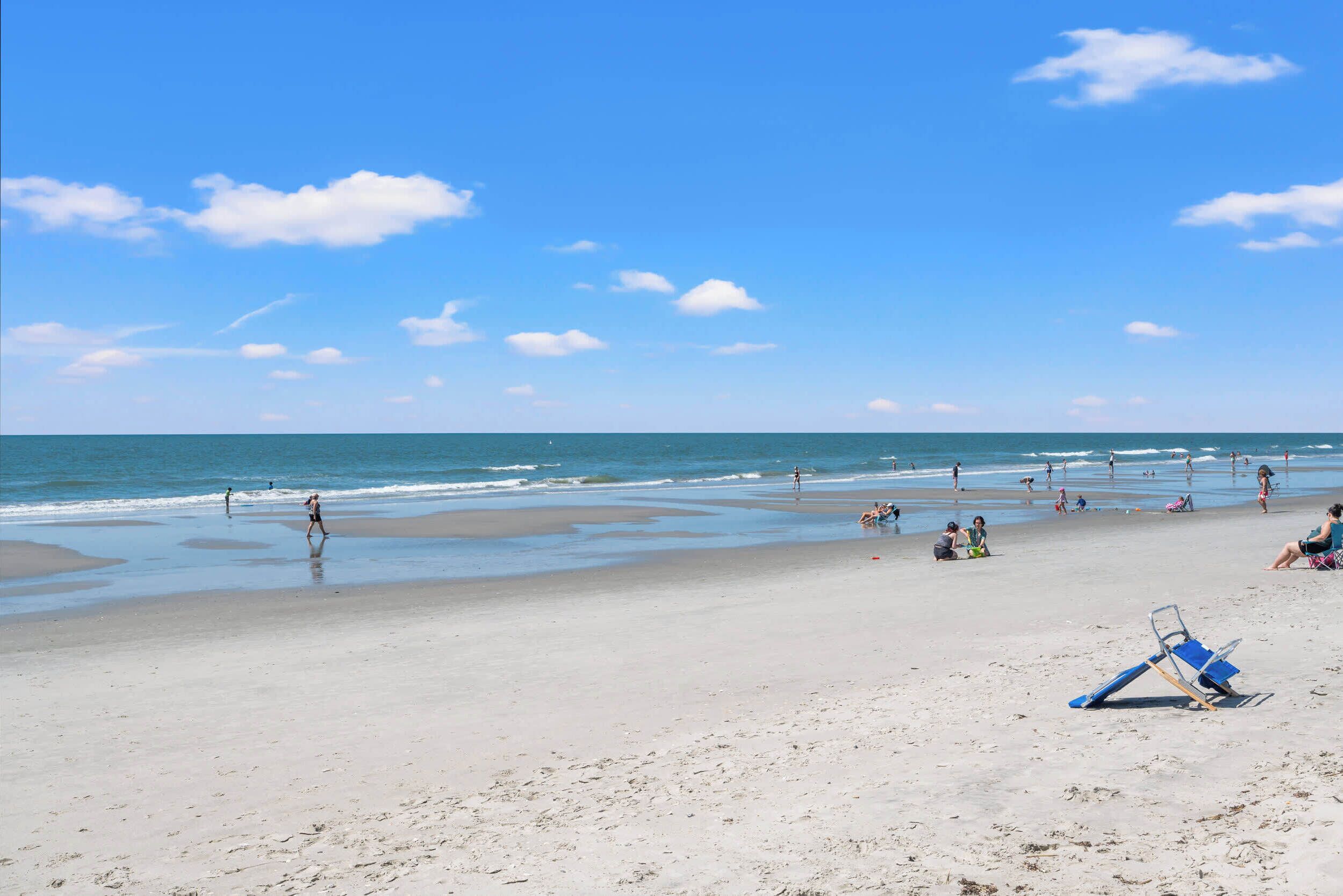A beach with people walking and sitting, a clear sky, and the ocean in the background.