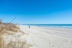 Ein weiter Sandstrand mit Fußabdrücken, einem klaren, blauen Himmel und einem Gebäude in der Ferne.