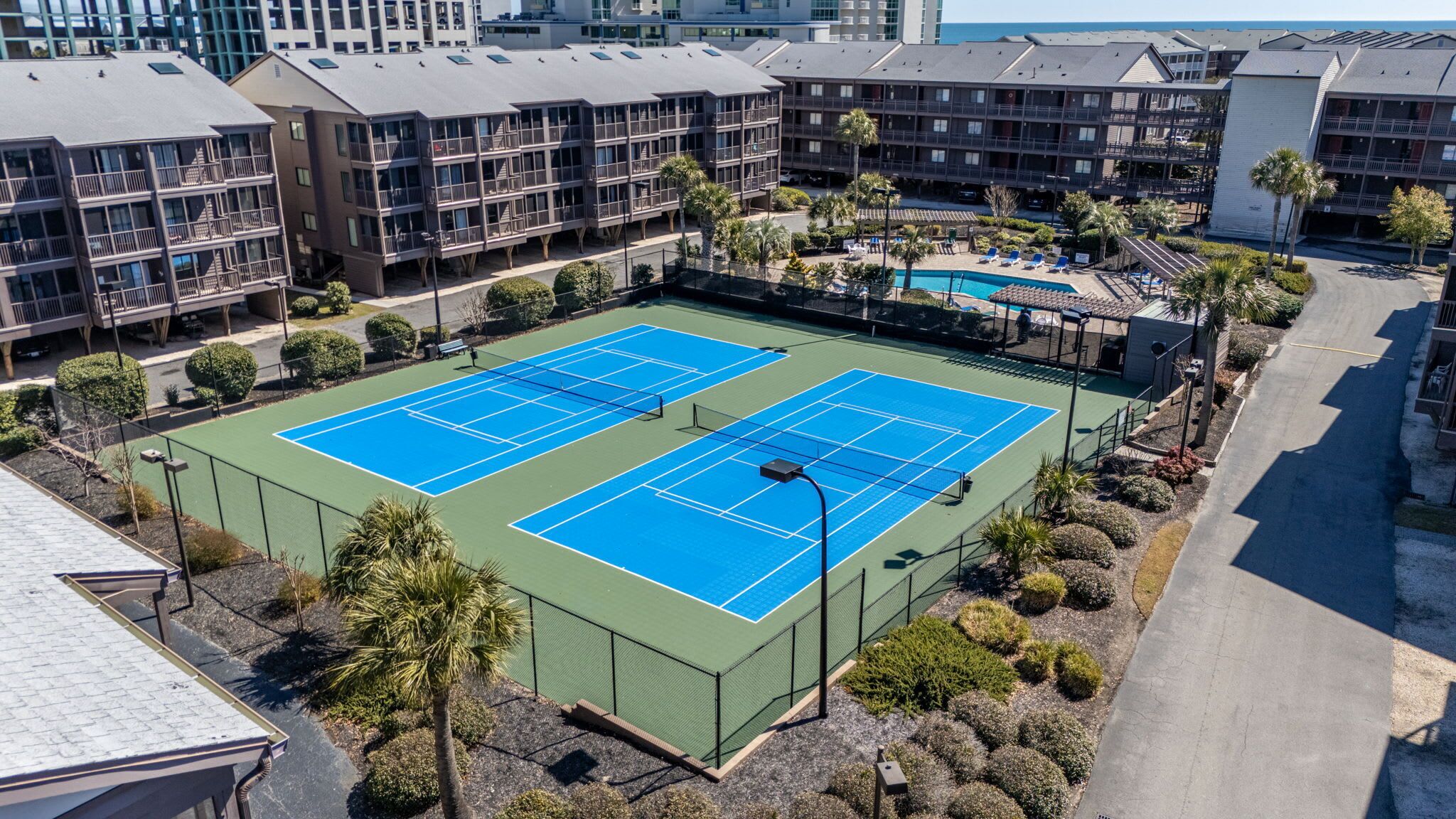 Un campo da tennis circondato da una recinzione, con una piscina e edifici sullo sfondo.