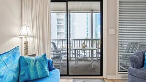 A room with a sofa, a lamp, a balcony with a table and chairs, and a window with blinds. - Orange Crush at Tilghman (North Myrtle Beach)