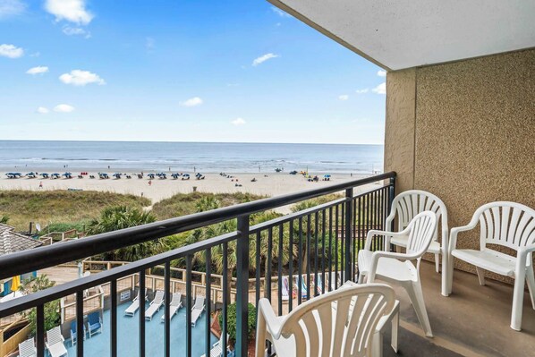 Un balcon avec vue sur la plage, des chaises en plastique blanches et un espace piscine.
