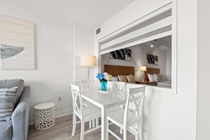 Une pièce moderne avec une table à manger blanche, des chaises, une petite table d’appoint ronde, un canapé et un miroir qui reflète une chambre à coucher avec un lit et une table de chevet.