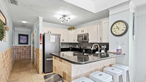 A kitchen with white cabinets, a black countertop, a stainless steel sink, a microwave, and a clock on the wall. - "The Pearl of Tilghman" (North Myrtle Beach)