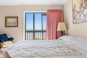 Een slaapkamer met een bed, een stoel, een lamp, een schilderij en uitzicht op het strand door een schuifdeur.