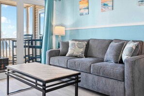 Une pièce avec un canapé gris, une table basse en bois, une lampe et une vue sur l’extérieur.