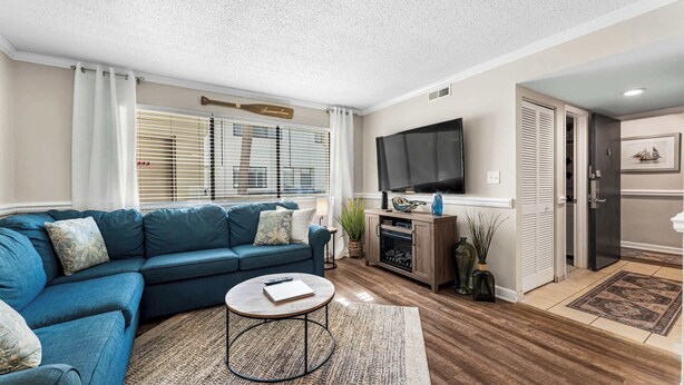 A living room with a blue sofa, a flat-screen TV, a small coffee table, and a kitchen area with a refrigerator and cabinets. - "Dreams By The Sea" at dunes village (Myrtle Beach)