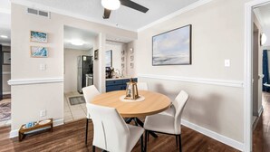 A dining area with a round table, white chairs, a ceiling fan, and a kitchen in the background. - "Dreams By The Sea" at dunes village (Myrtle Beach)