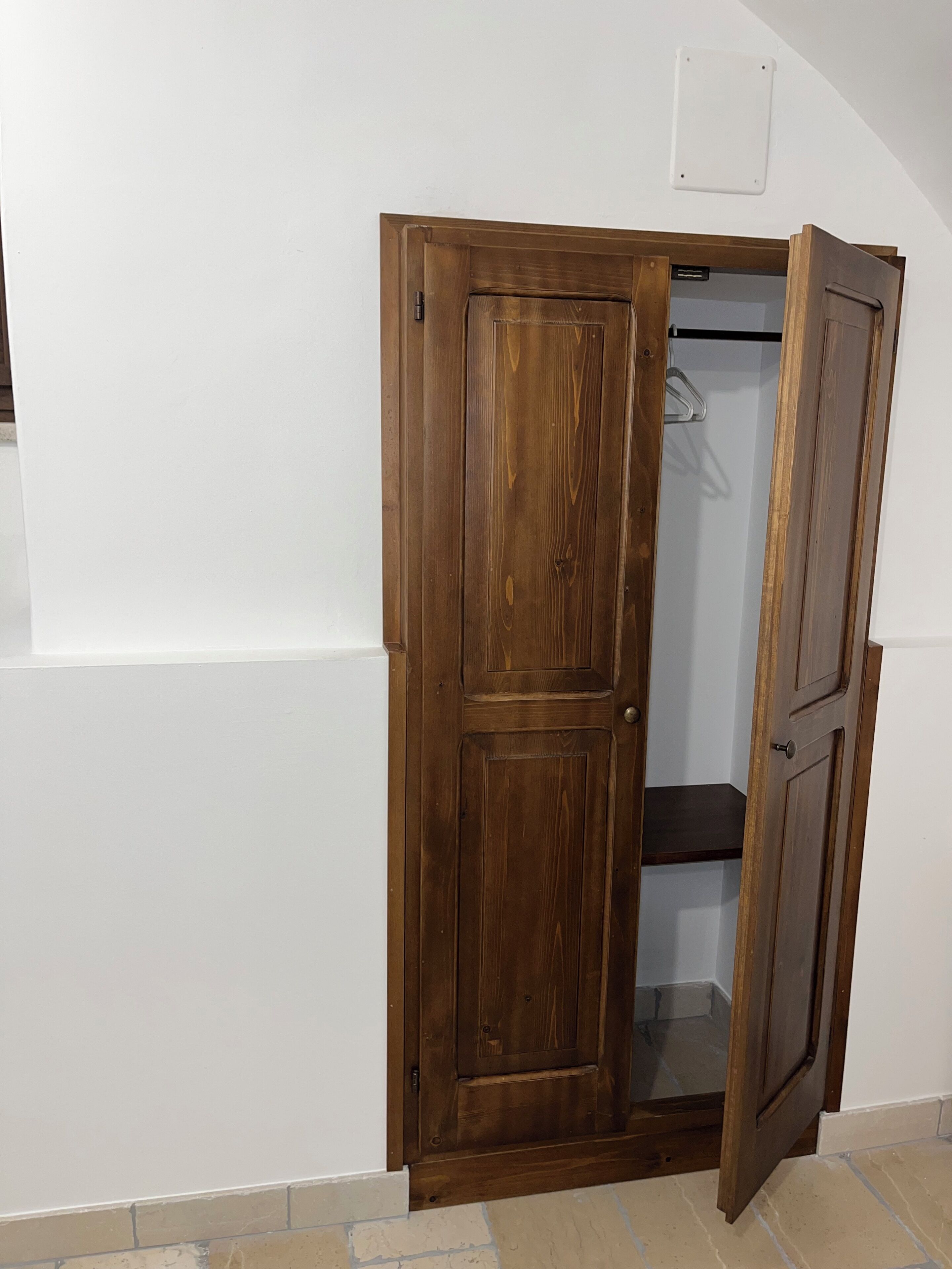 Une armoire en bois, dotée d’un miroir et d’une tringle pour suspendre les vêtements, est placée contre un mur blanc, au-dessus d’un sol carrelé.
