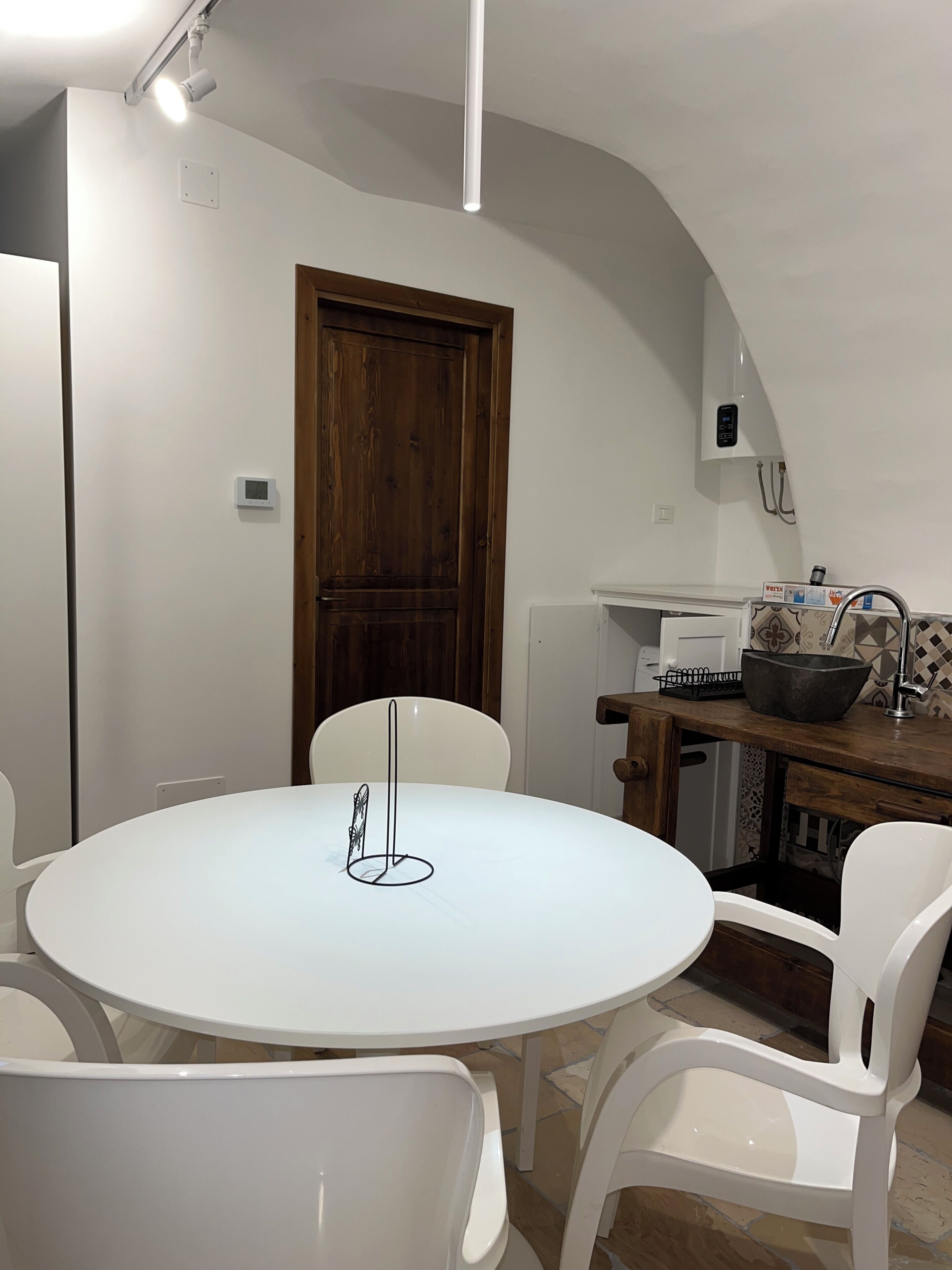 A dining area with a round white table, white chairs, a wooden door, and a kitchen area with a sink and a large bowl.