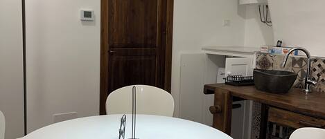 A dining area with a round white table, white chairs, a wooden door, and a kitchen area with a sink and a large bowl.