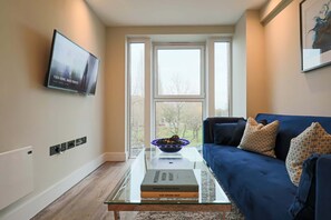 A modern living room with a glass coffee table, a blue sofa, and a large window with a view of trees. - 🌟Stylish 1 Bedroom Apartment Central Welwyn Garden City -Ideal for Family Visit (Welwyn Garden City)
