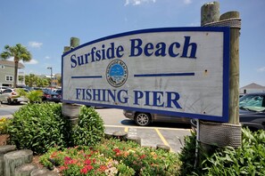 Señal del muelle de pesca de Surfside Beach, con vista clara del estacionamiento y la vegetación circundante.