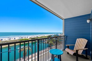 A balcony with a view of the beach, a chair, and a small table.