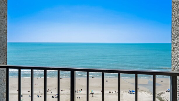 Uitzicht op een strand vanaf een balkon met een hekwerk, een zandstrand en een rustige zee.