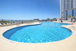 A large outdoor swimming pool with a sandy beach area, surrounded by a fence and a tall building in the background.