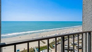 Uitzicht op een strand vanaf een balkon, met een heldere lucht, zandstrand en geparkeerde voertuigen.