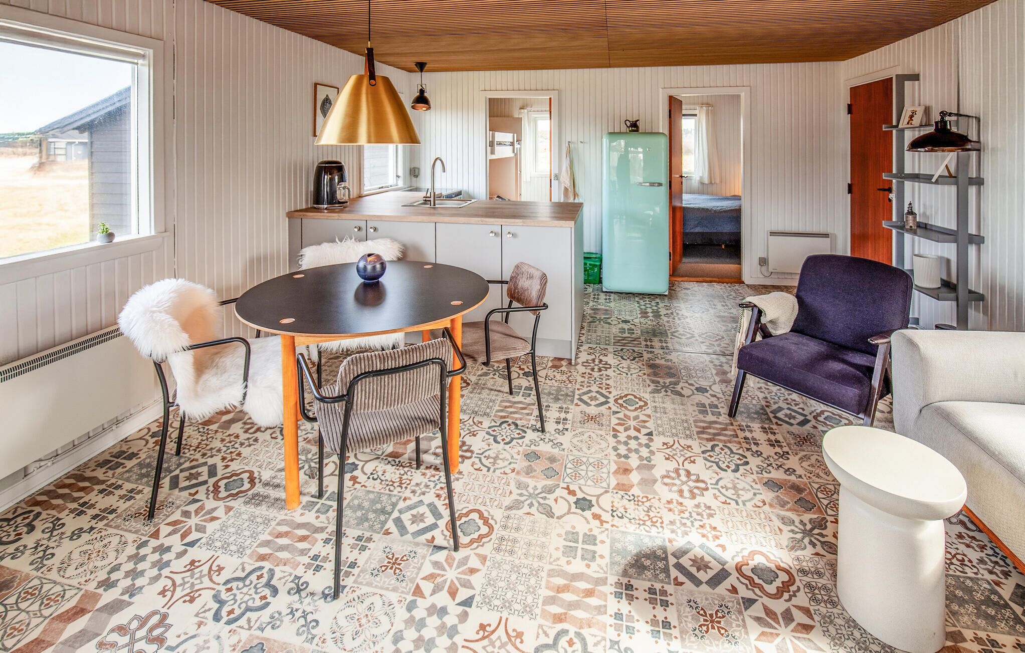 A cozy interior with a round table, chairs, a sofa, a kitchen area, and a patterned floor.