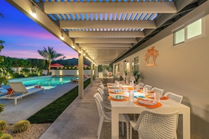 An outdoor dining area with a pool, white chairs, and a table set with orange placemats. - Living The Desert Way - Expansive 5‑Bedroom with Pool, Spa, Fire Pit & Stunning Mountain Views (Palm Springs)