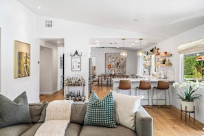 A modern living room with a sofa, a dining area, and a kitchen. - Living The Desert Way - Expansive 5‑Bedroom with Pool, Spa, Fire Pit & Stunning Mountain Views (Palm Springs)