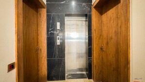 A hallway with a wooden door, a black marble wall, and a glass elevator door. - Shri Ram Retreat (Vrindavan)
