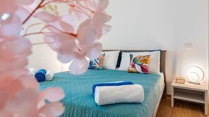 Un letto rifatto con cura, con copriletto color petrolio, un asciugamano bianco e cuscini colorati. È presente un comodino con una lampada e uno specchio.