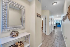 A hallway with a decorative mirror, a welcome sign, a basket on a console table, and a tiled floor. - Legacy Tower II 206 | Stunning Coastal Views! (Gulfport)