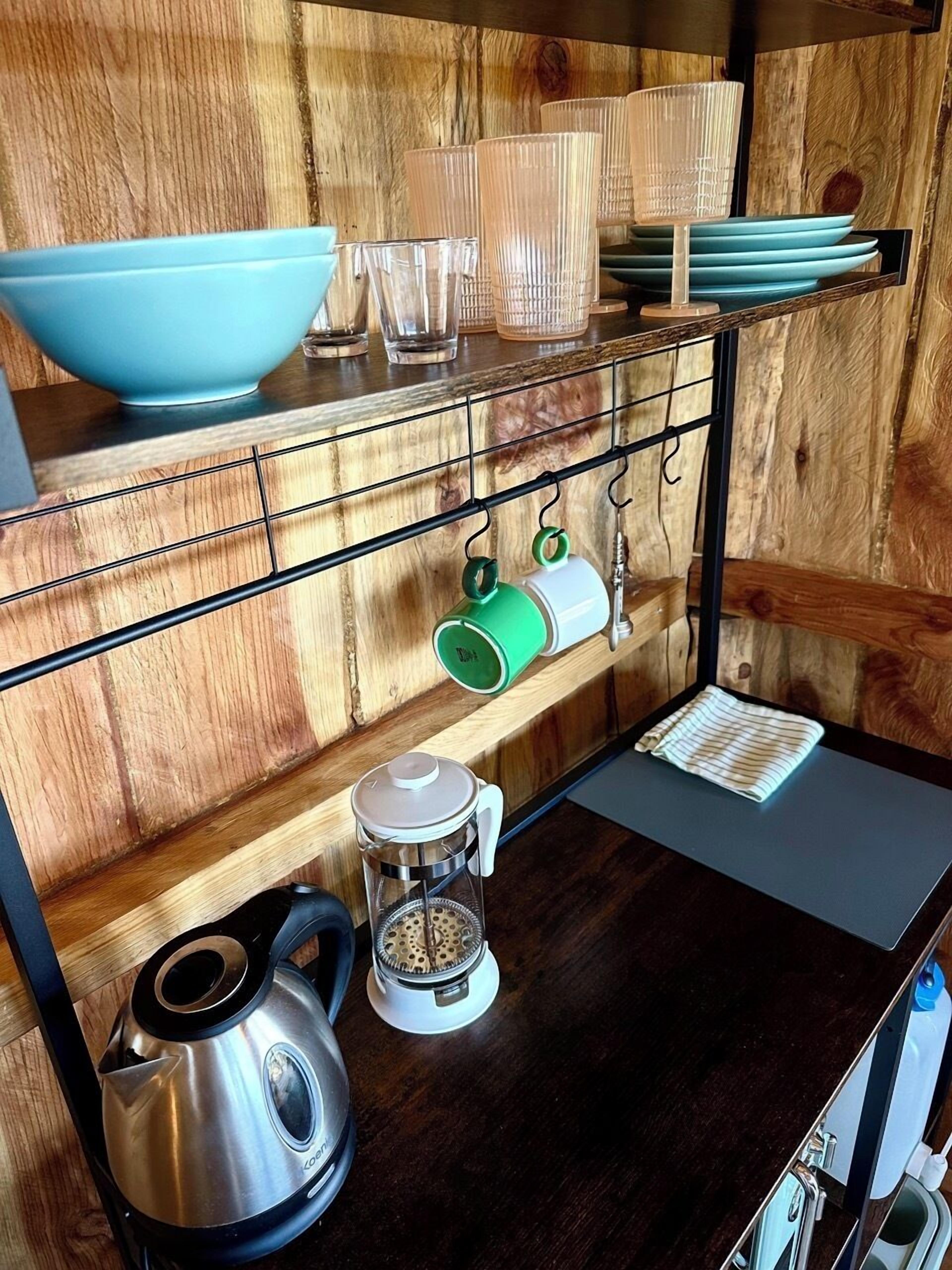 A kitchen with a wooden wall, a metal rack holding cups and bowls, a kettle, and a coffee maker.