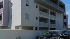 Un edifici modern d'àmbit diversos amb balcons, teulada plana i zona d'aparcament amb cotxes.