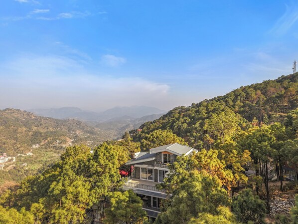 A modern building with multiple floors, surrounded by dense forest and hills.