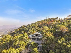 A two-story building with a traditional architectural style, surrounded by dense forest and hills in the background. - ELIVAAS 4 BHK Kavya Villa With Lounge  (Kasauli)