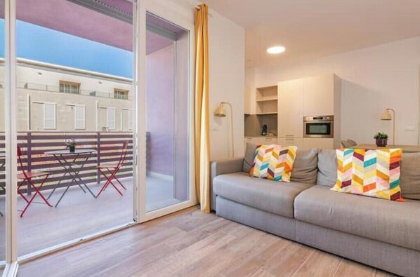 A modern living room with a sofa, colorful pillows, a dining area with a table and chairs, and a kitchen area with a microwave. - Apartment with Balcony - 900 m from Tower, Private Park & Modern Comfort (Pisa)