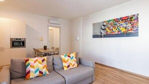 Un salon moderne avec un canapé gris, des coussins colorés, un coin repas comprenant une table et des chaises, et une peinture abstraite aux couleurs vives accrochée au mur.