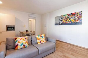 A modern living room with a grey sofa, colorful cushions, a dining area with a table and chairs, and a vibrant abstract painting on the wall. - Apartment with Balcony - 900 m from Tower, Private Park & Modern Comfort (Pisa)