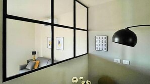 Ein Zimmer mit einem großen Spiegel, einer schwarzen Schreibtischlampe, einem kleinen Tisch mit Blumen und einem an Wand montierten Schild.