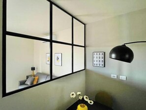 A room with a large mirror, a black desk lamp, a small table with a vase of flowers, and a wall with a framed sign. - New Apartment with Balcony - 900 m from the Tower, Private Park (Pisa)