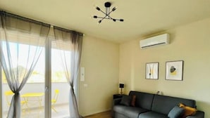 A living room with a sofa, a table, and a chair. There is a ceiling light fixture and an air conditioning unit on the wall. - New Apartment with Balcony - Near Tower, Private Park (Pisa)