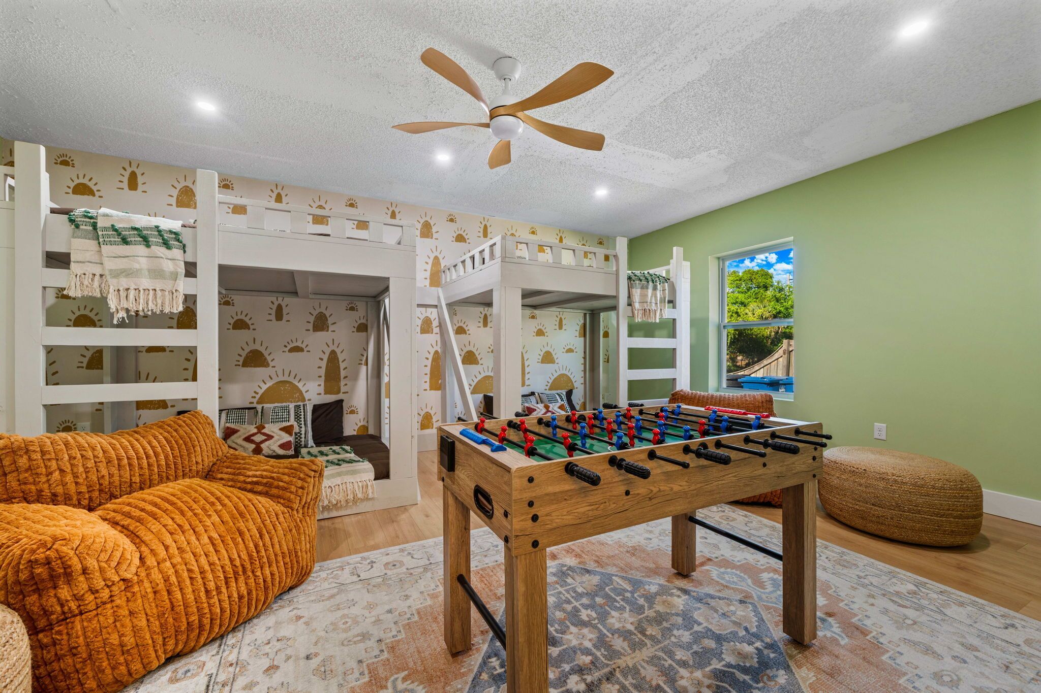 A room with bunk beds, a foosball table, a ceiling fan, and a window with a view.