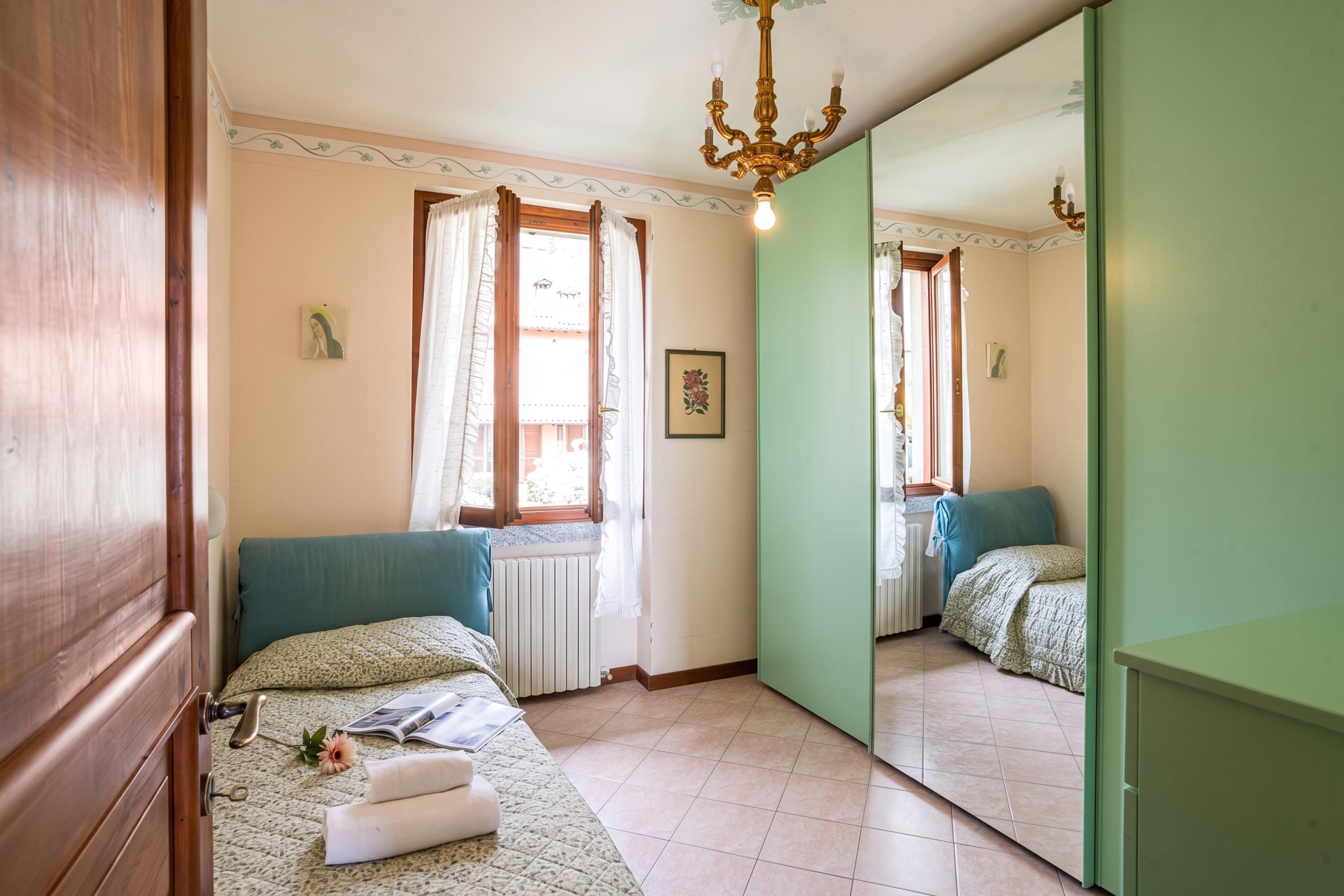 A bedroom with a bed, a window with curtains, a radiator, a mirror, and a chandelier.